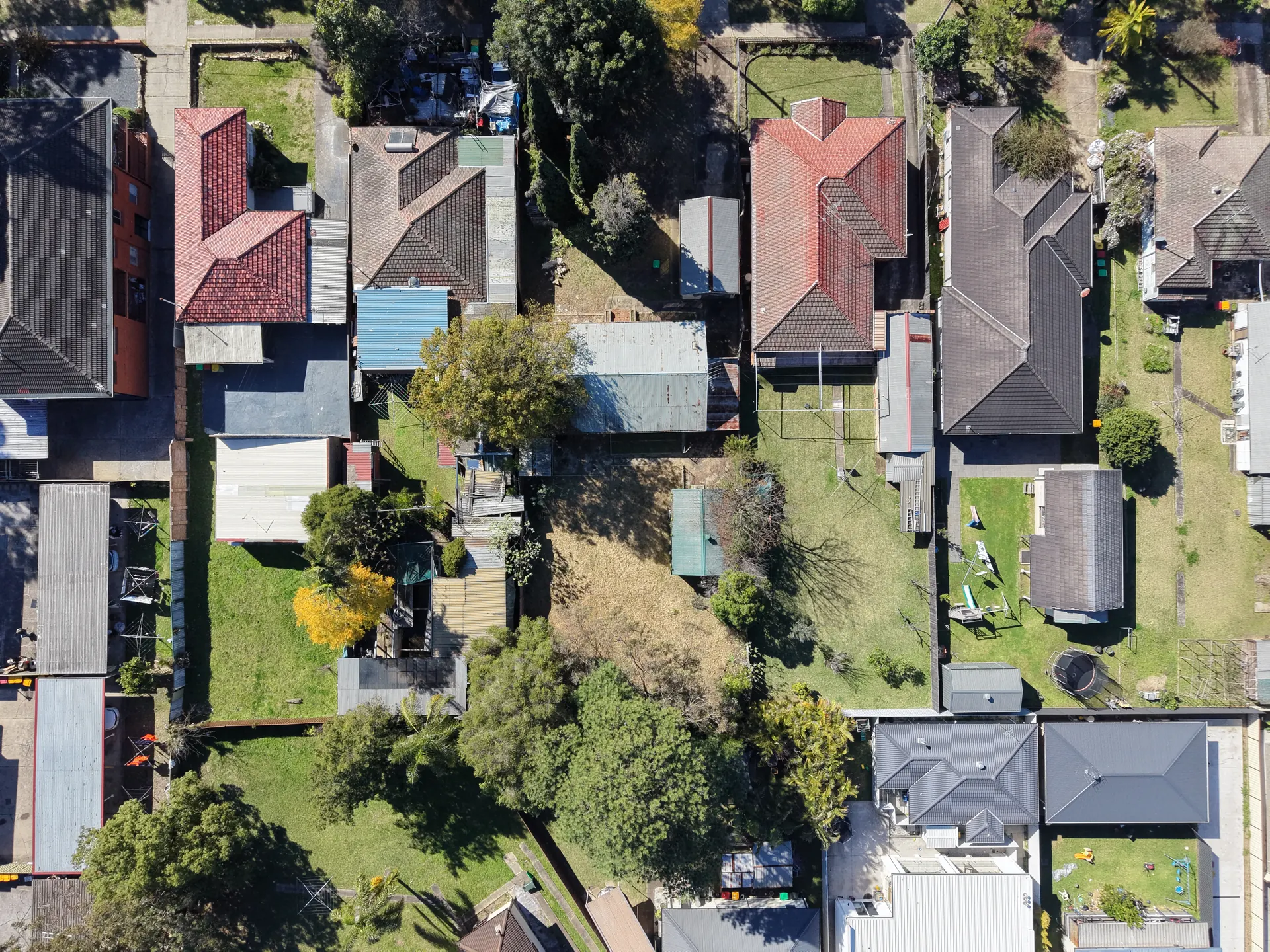Aerial view down onto a suburban block, with a long narrow building and its bare yard sitting between neighbouring houses, sheds and gardens.