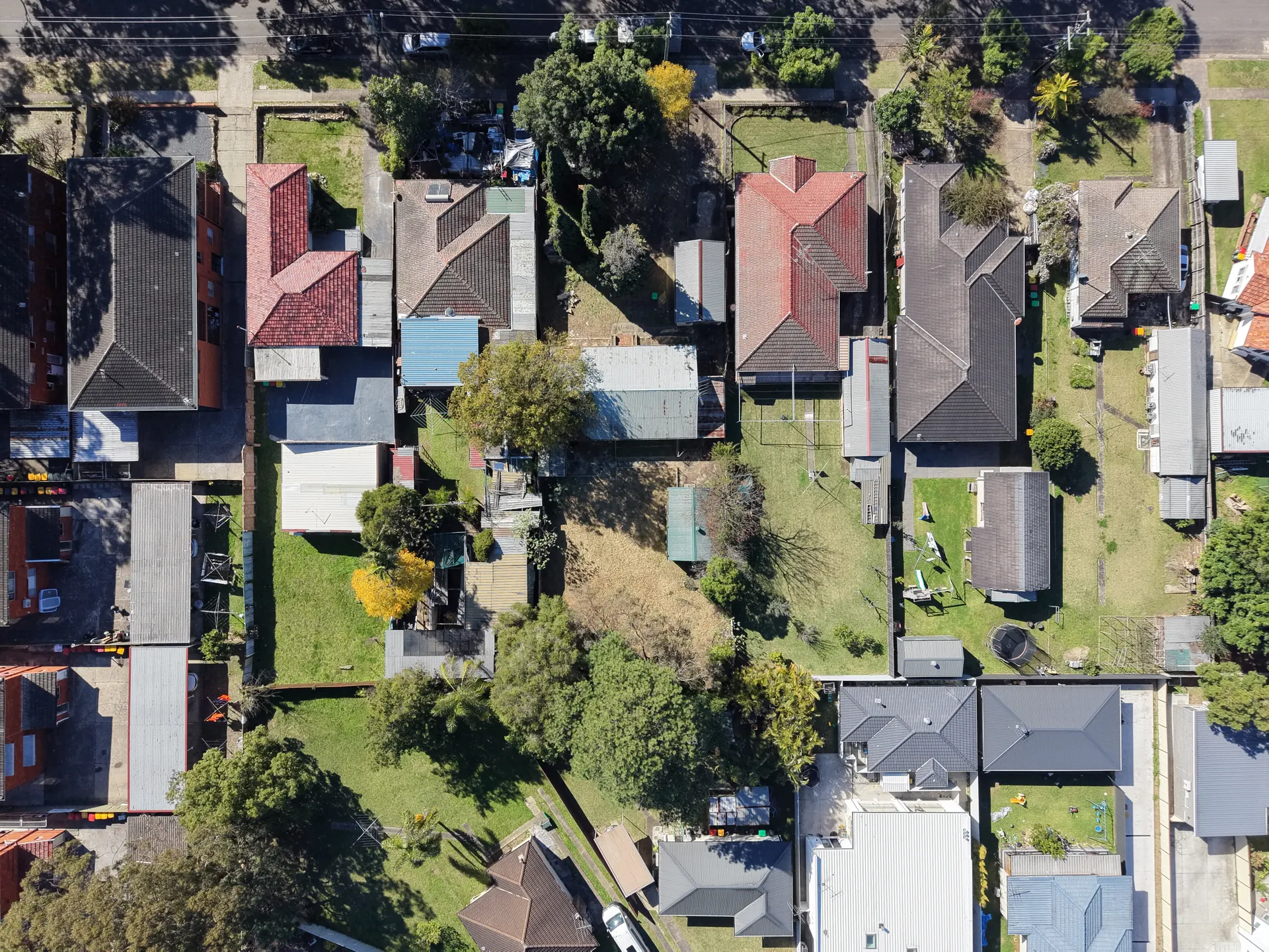 Aerial view looking straight down over a residential neighbourhood: two roads, around twenty-five single-storey houses with tiled and metal roofs, fenced backyards, garden sheds and mature trees.