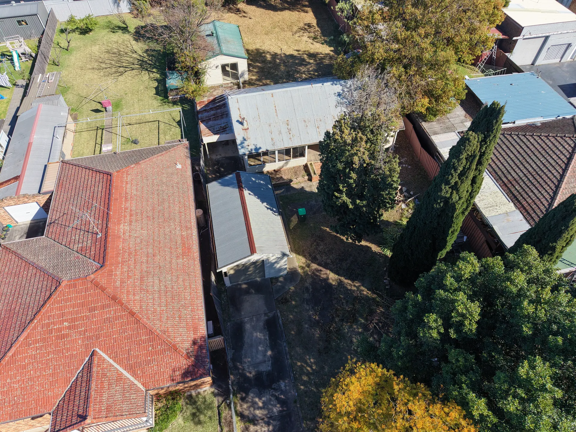 Low aerial view of the building and its yard, with the roofs of neighbouring houses close on either side and trees along the rear boundary.