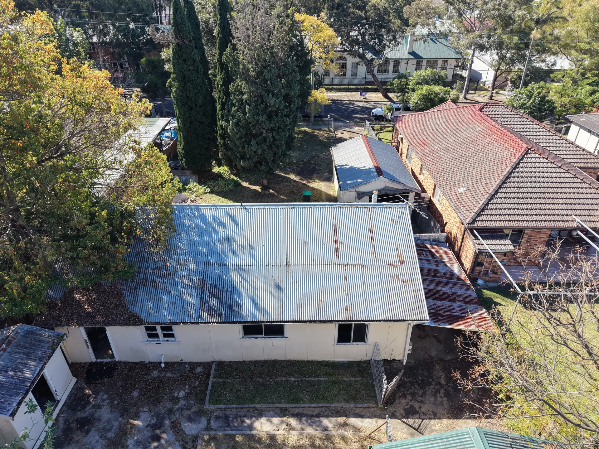 Aerial view of a single-storey building with a rusting corrugated-iron roof on a long suburban block, with tall cypress trees on one side, neighbouring houses close by and a street beyond.
