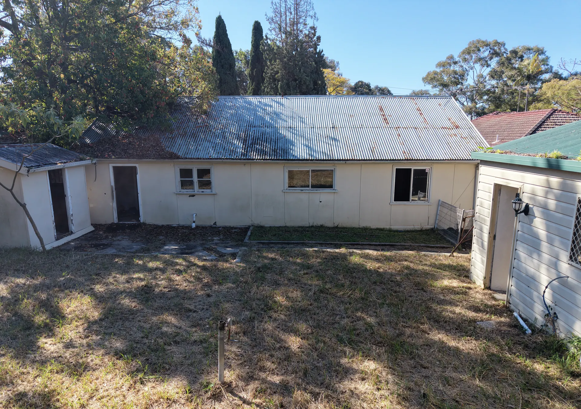 Ground-level view of the rear of the single-storey building: a rusting corrugated-iron roof, rendered walls, one window standing open to the weather, a doorway with no door, and a dry unlandscaped yard between two outbuildings.