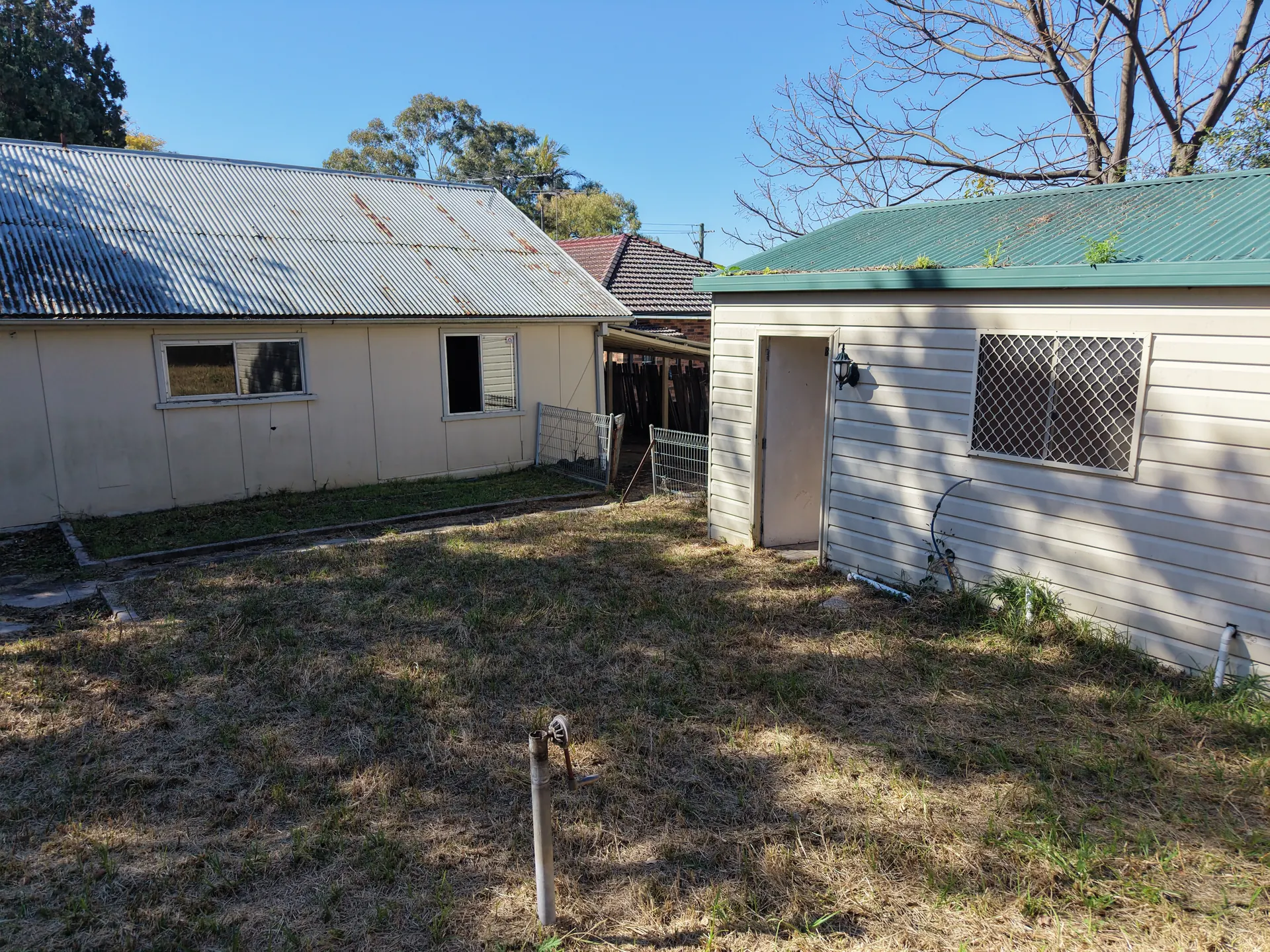 Ground-level view along the building's rear wall and open doorway, across a dry unlandscaped yard, with a clad outbuilding standing alongside.