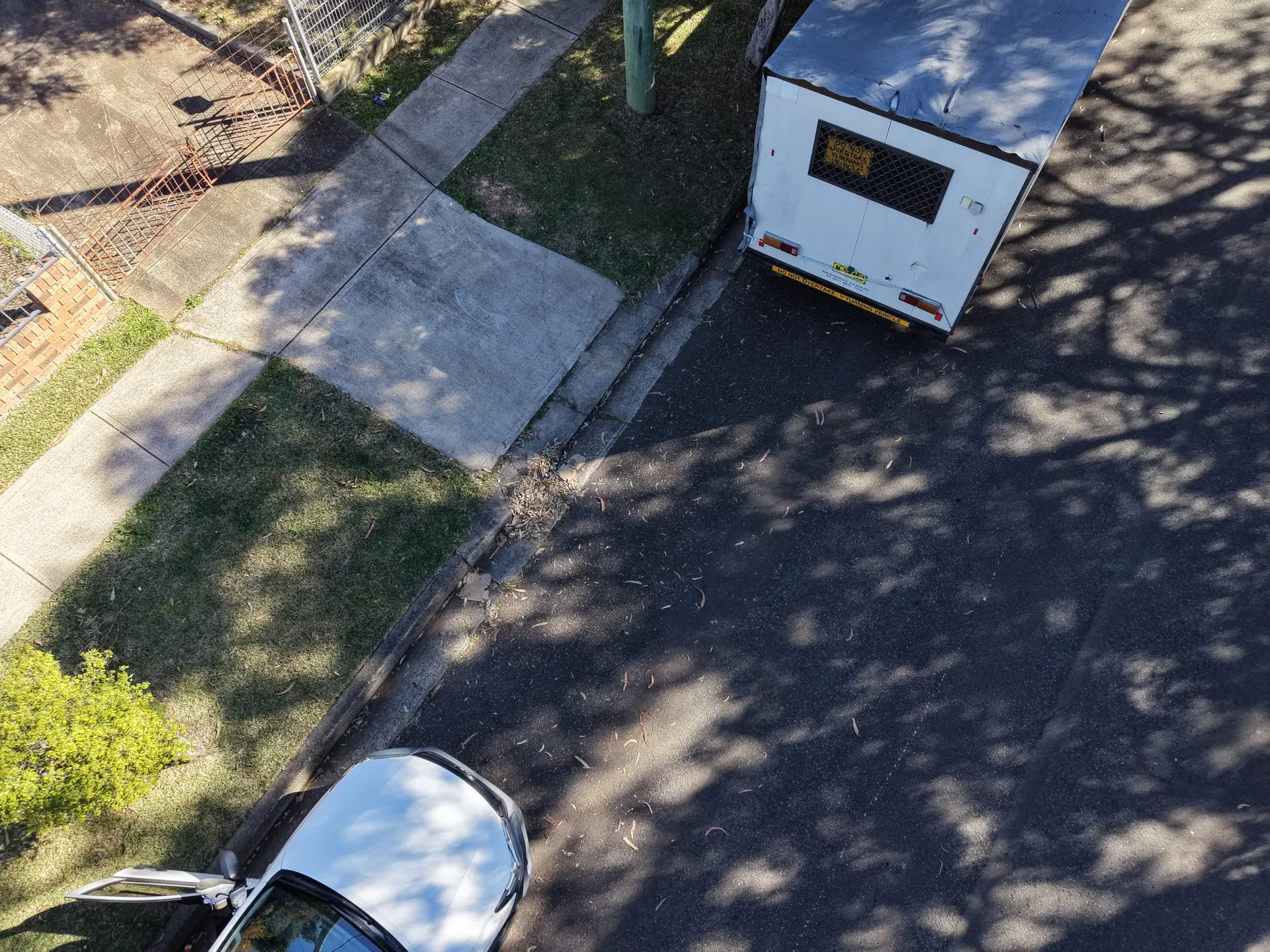 Low overhead view of a suburban kerb, concrete footpath and grass verge, with a power pole, a parked box trailer and the roof of a car at the roadside.